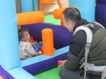 お父さんが見守っている横のボールプールで遊んでいる幼い子の写真