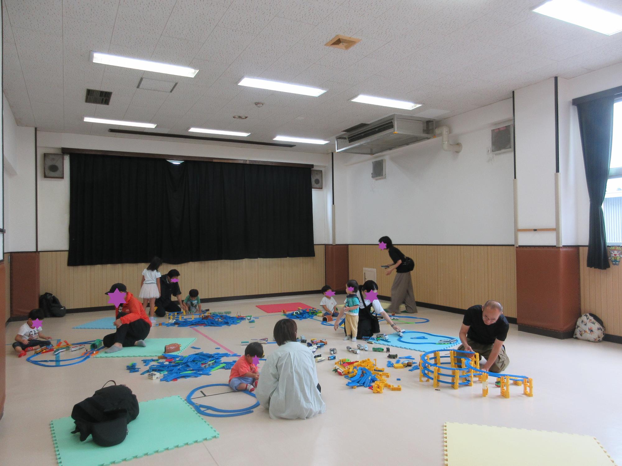 集会室に広がったプラレールを5組の親子がそれぞれ好きな形に組立て遊んでいるところ