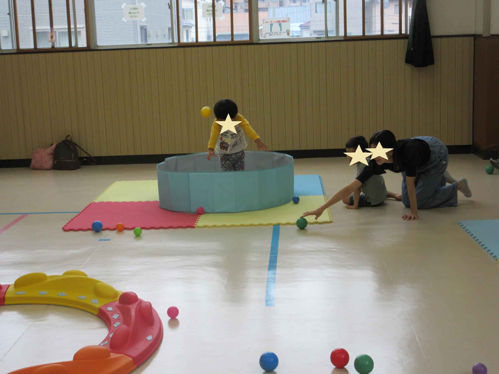 幼児がボールプールに入り投げられたボールに肩をすくめ受け止める幼児と床に転がるボールをつかむ母とみている兄