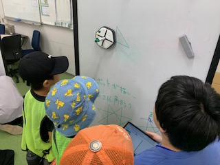 児童が、壁に張り付いているロボットをプログラミングして図形を描いている様子の写真