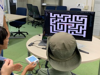 大学生が作製した、コントローラーを実際に動かすジャイロセンサーを用いたゲームを体験している児童の様子の写真