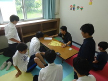 1人の男子高校生がどきどきトランプに挑戦する周りで観戦する高校生と小学生の写真