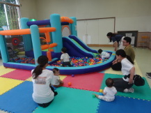 幼児たちが楽しそうに遊具で遊ぶ姿を外側に座って見守っている親たちの写真