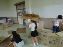 軍手を付けて頭に乗せながら段ボールを運ぶ女の子と周りで段ボールハウスを作る子供たちの写真