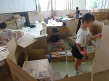 座った子供が隠れるくらいの段ボールの壁を作っていく3人の子供たちの写真