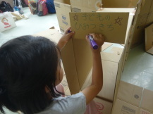 段ボールハウスの入り口に看板を書いている女の子を撮影している写真