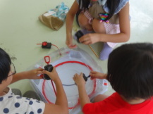 ベイブレードスタジアムの台の周りを円状になって座った3名の子どもたちがそれぞれベイブレードを持っている写真