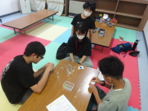 たくさんのクリップが置かれたテーブルの上で2名の子どもがクリップつなぎに挑戦している様子を2名の子どもが見守っている写真