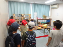 子どもが赤い境界線が設置されたテープの前に立ち、遠くのおもちゃに向けて射的を行っている様子の写真