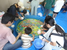 水がはられたくさんのスーパーボールが浮かんでいる家庭用子どもプールの周りを親子が数組が座り、スーパーボールすくいを楽しんでいる写真