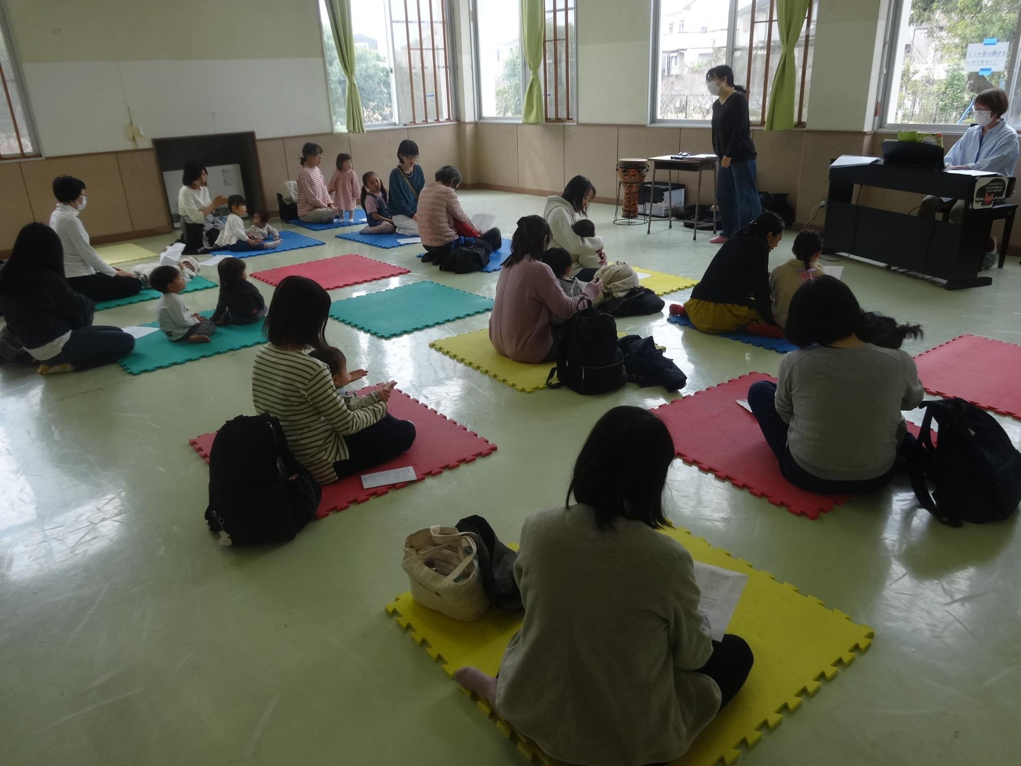 トレーニング室にカラーマットを並べ、乳幼児親子が座って先生の話しを聞いている写真
