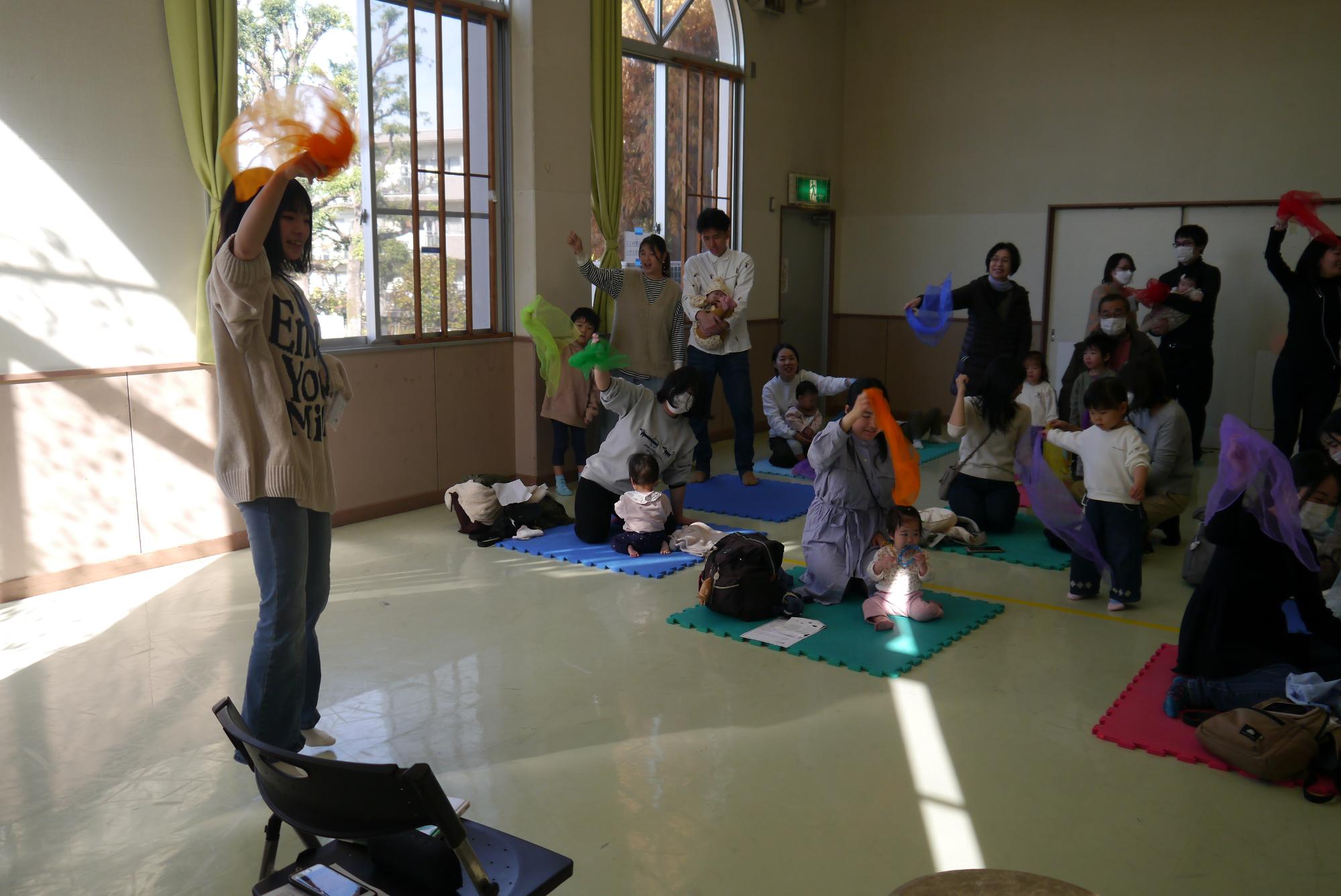 「できっこないをやらなくちゃ」の曲にのせてスカーフを持って踊る乳幼児親子とボランティアの高校生の写真