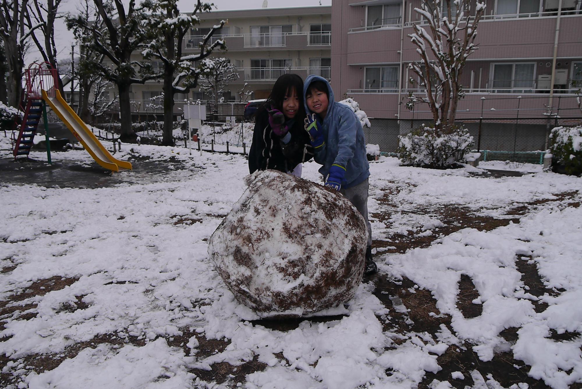 大きな雪玉を作ってピースサインをする小学生の姉弟の写真