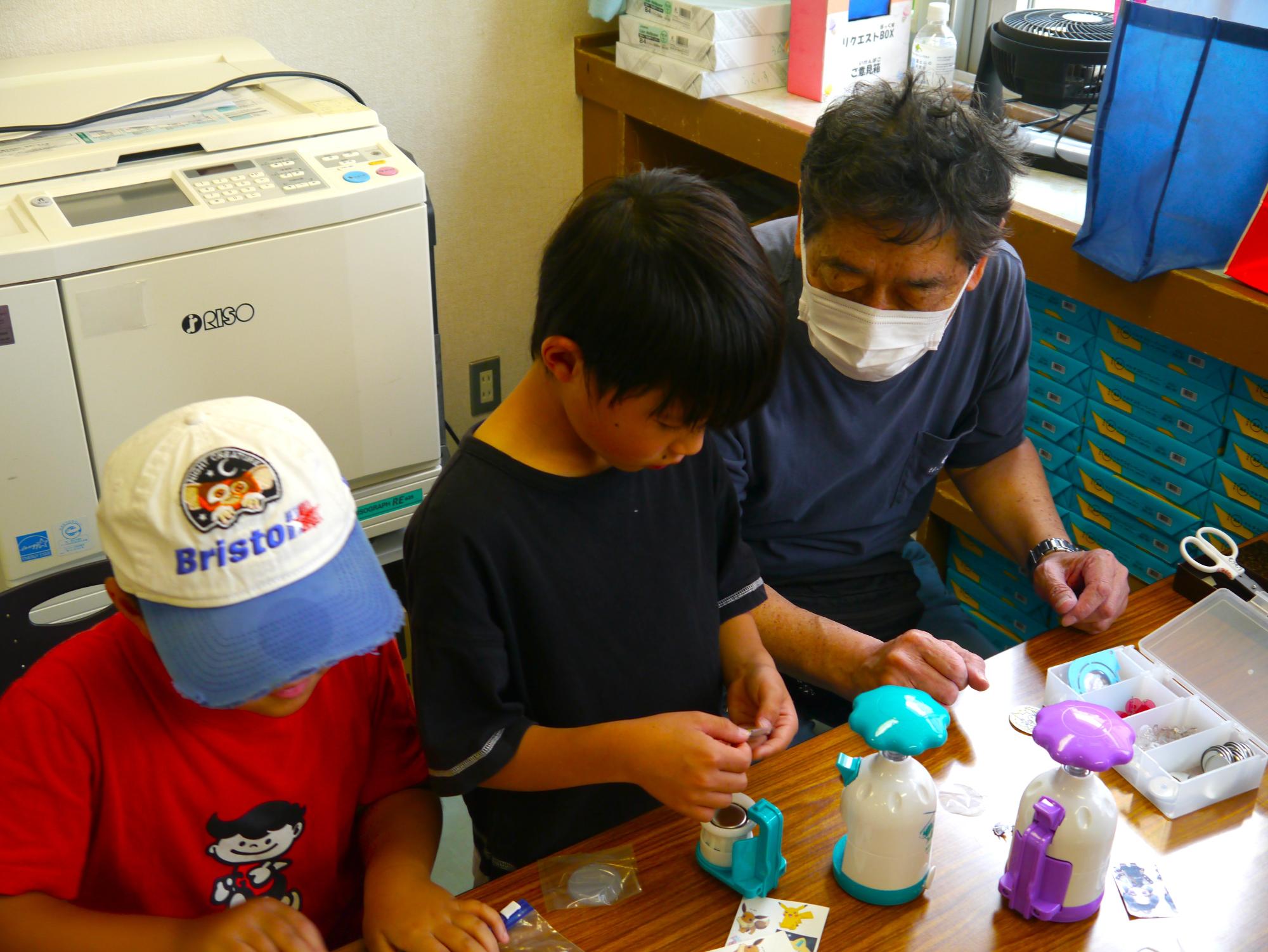 缶バッジマシーンでイラストを金具に取り付ける作業をする小学生と先生