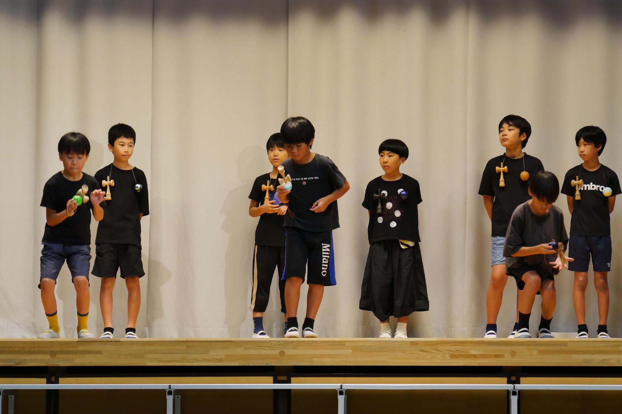 体育館のステージの上でけん玉の技を披露する3人の小学生の写真