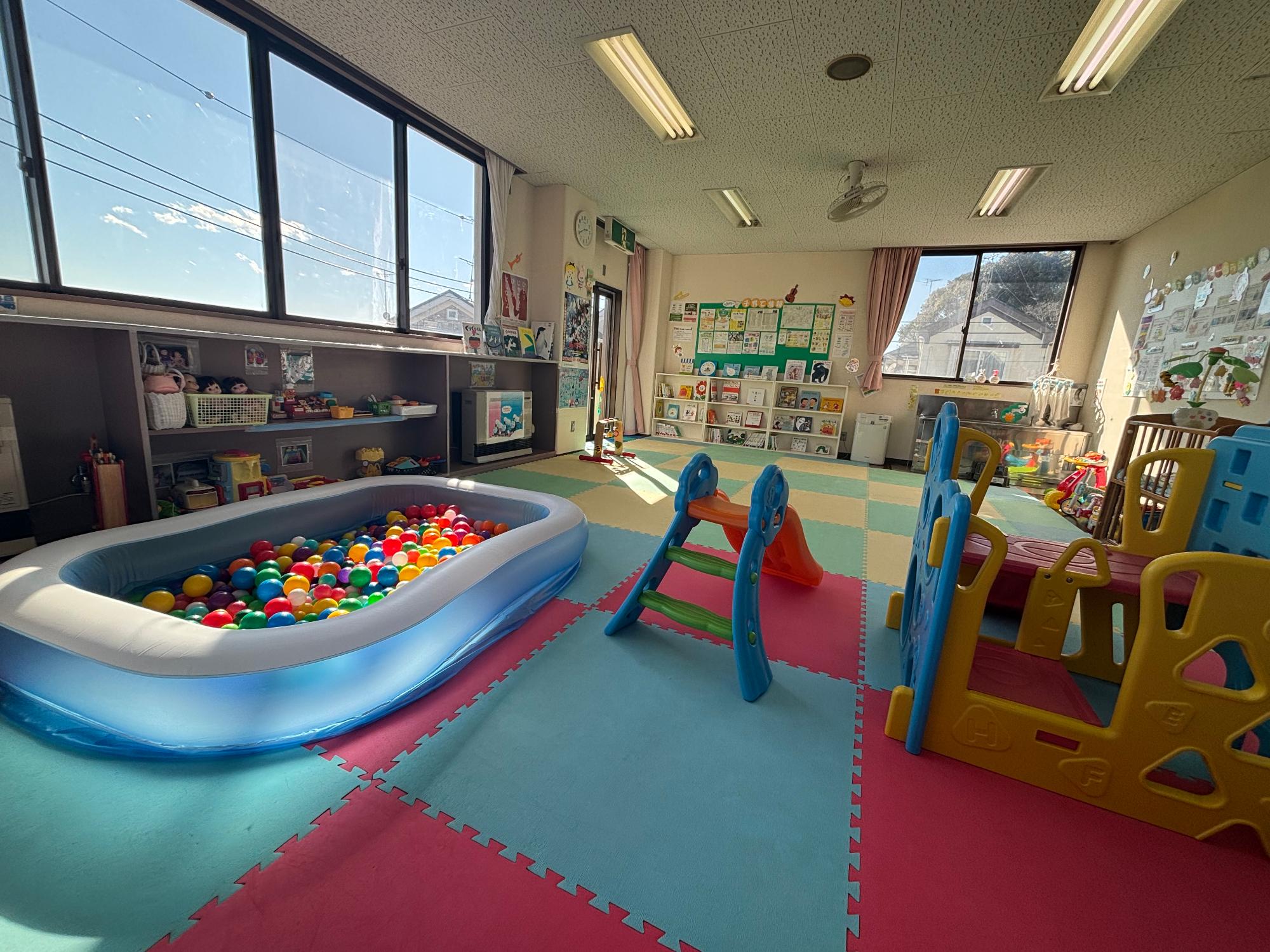 カラフルなマットの上に滑り台やおもちゃがあり、棚に絵本が並べられた幼児室の写真