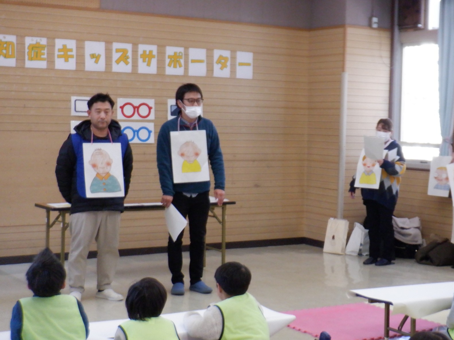 職員が芝居をしながら読み聞かせをしているのを小学生児童が聞いている様子の写真