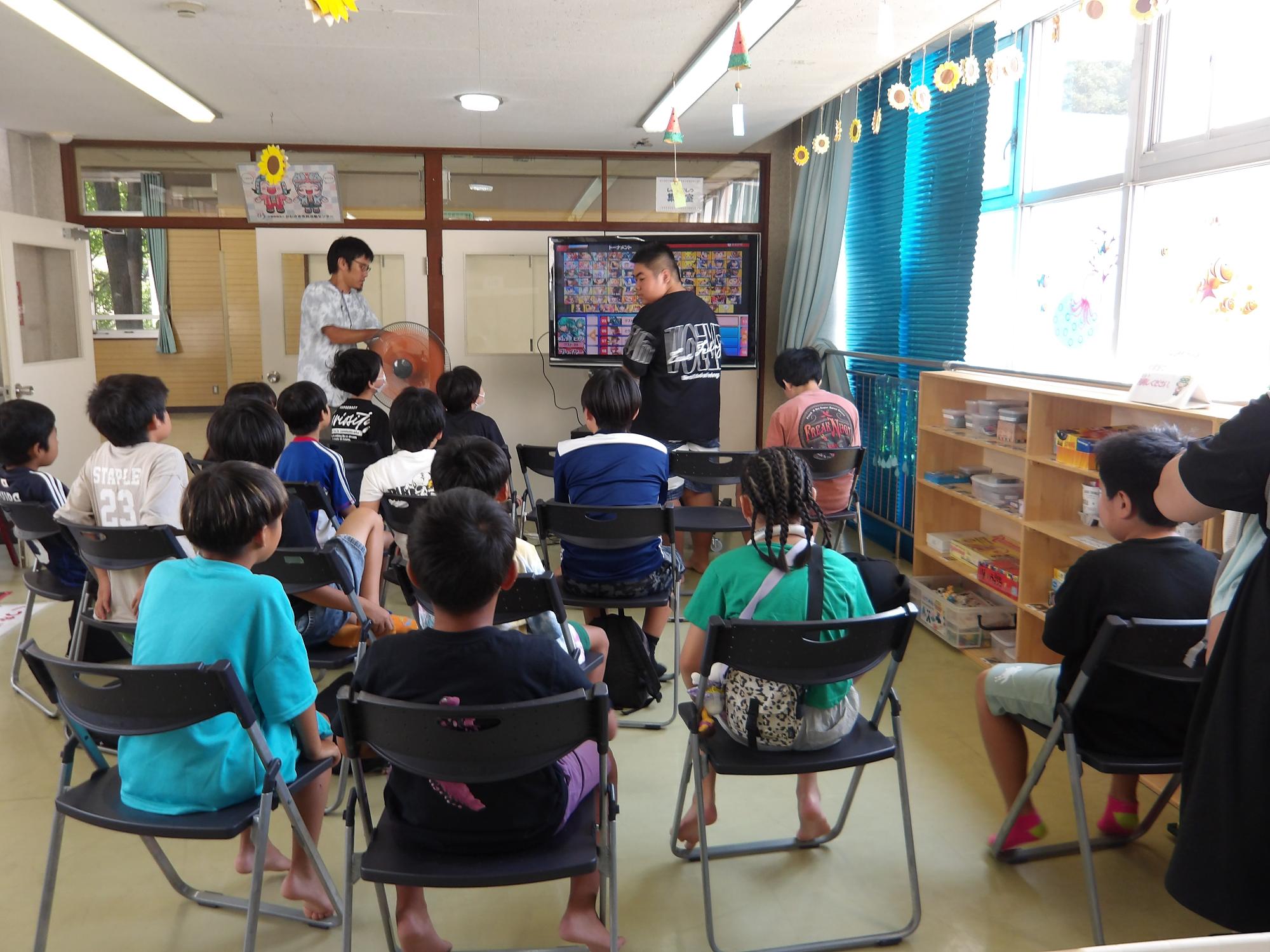 ひまわりの絵などで装飾された室内で大きなテレビモニターの前に複数の子どもたちが椅子に座りゲームを楽しんでいる様子の写真