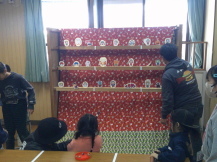 三段の棚に各列8個ほど置かれた射的の的を狙う親子と順番を待つ2人の女の子の写真