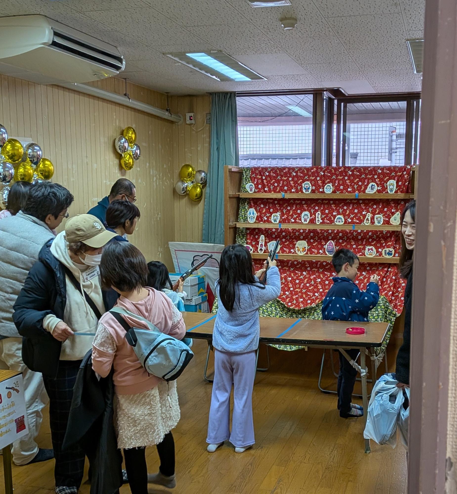 三段の棚に各列8個ほど置かれた射的の的を狙う親子と順番を待つ2人の女の子の写真