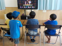 ゲームで対戦をしている3人の男の子の後ろで観戦する青い帽子を被った男の子の写真
