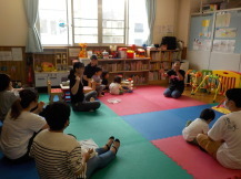 本が沢山置いている部屋の隅に座り読み聞かせをする男性と座って聞く親子たちの写真