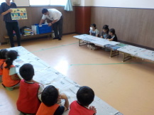子供たちが長机に座って、前で立っている大人が持っているイラストを見ている写真