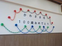 壁に飾られた「かわさき市100歳おめでとう」の文字の周りをペーパーフラワーと輪飾りで飾られている写真
