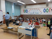 ホワイトボードの前に立つ男性の方に子供たちが一斉に指差しをしている写真