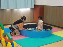 たくさんの小さいボールが入っている円形のプールに幼児が立って、それを見ている保護者の写真