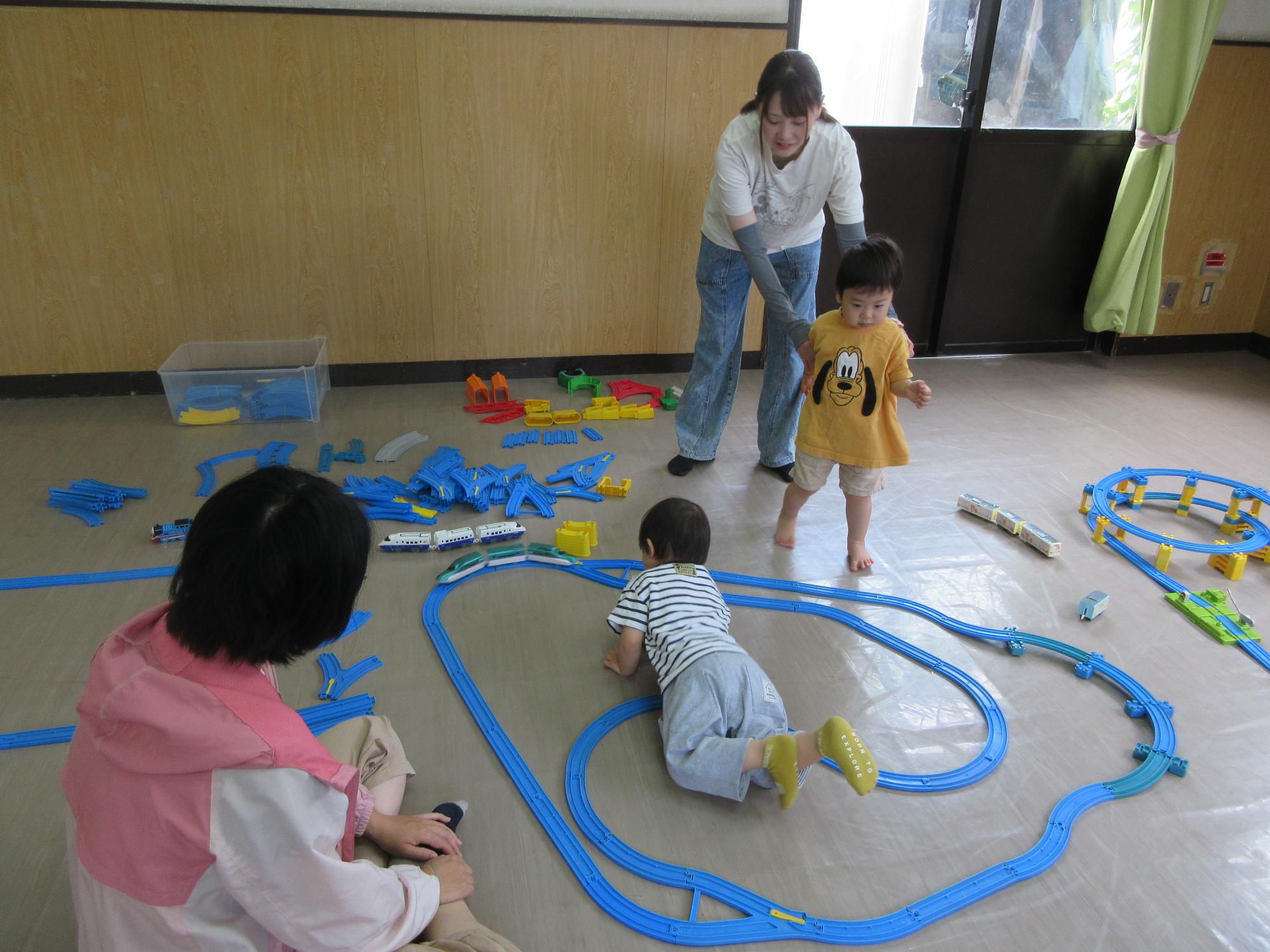 自分で作ったレイアウトでプラレールを走らせて遊ぶ幼児と保護者の写真