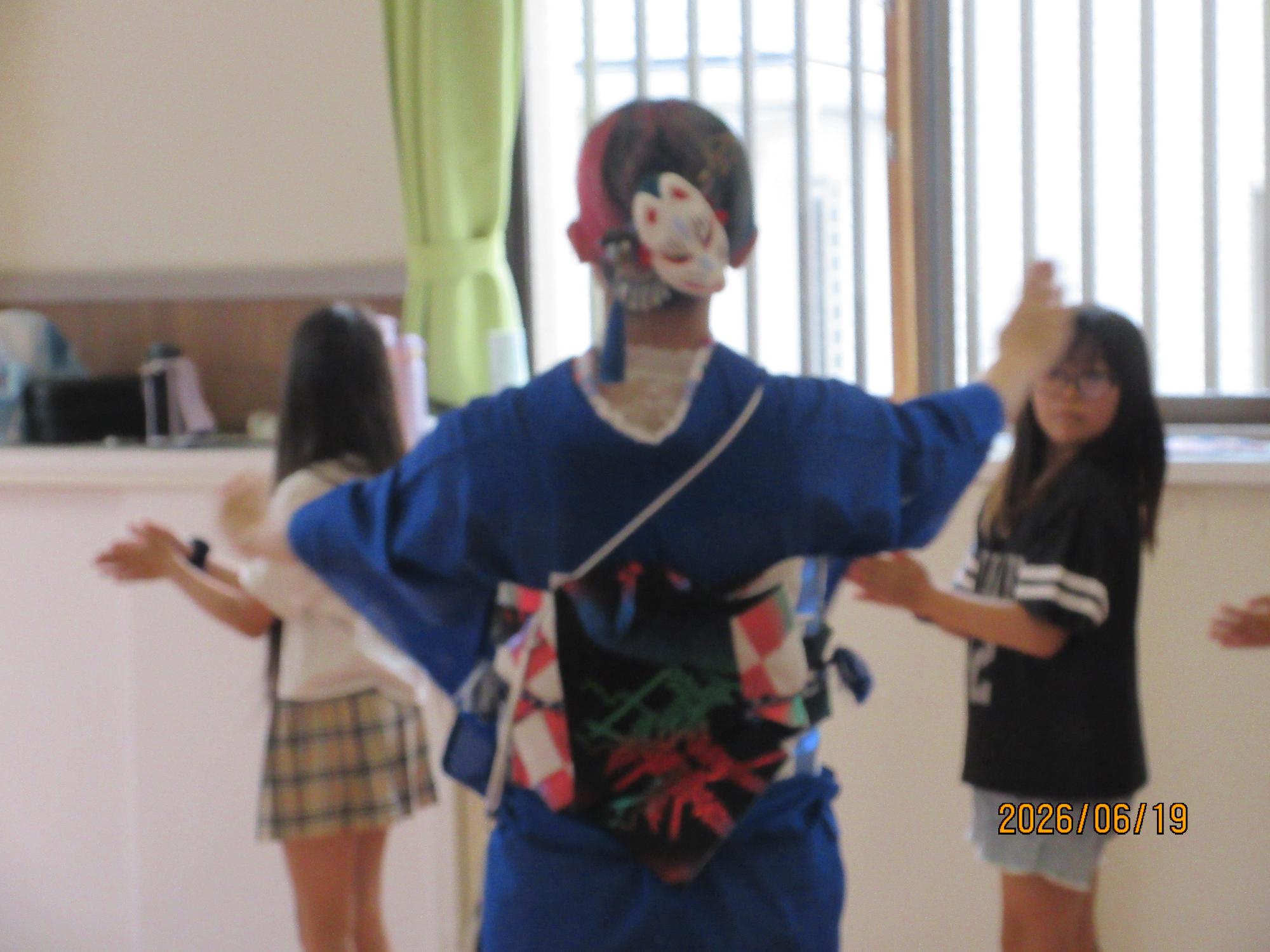 子どもたちが集会室で講師の先生と盆踊りを踊っているところの写真