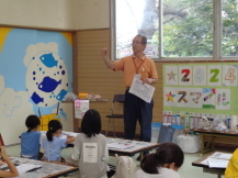 長テーブルが等間隔に設置された部屋で、子どもたちが正面に立つ先生の方を向いて話を聞いている様子の写真