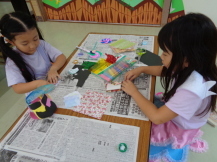 長机に置かれた新聞紙の上で、折り紙やセロファンを使い、工作をしている2名の女の子を写した写真