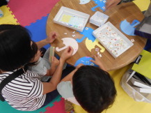 お母さんの膝に乗りながらシールを貼っていく幼児と横で座って作業する子供の写真