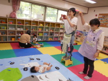 マットで仰向けの乳幼児をおもちゃを使い気を引かせる職員と隣でカメラを構える母親の写真