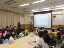 資料が映し出されたプロジェクタースクリーンを見つめる子どもたちと、スクリーンの近くに立っている関係者の写真