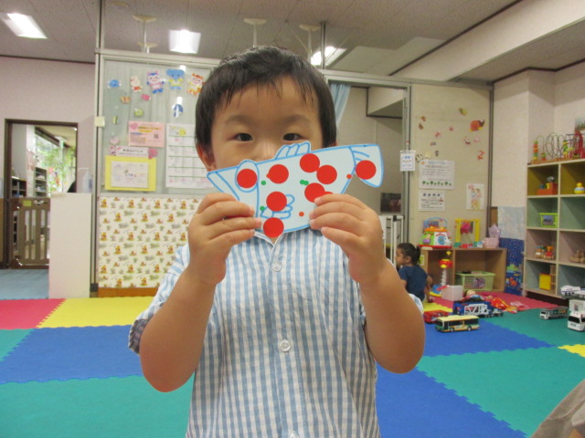 画用紙に描かれたさかなに赤や緑の丸シールを貼り、目の前で見せるようにもっている幼児の写真