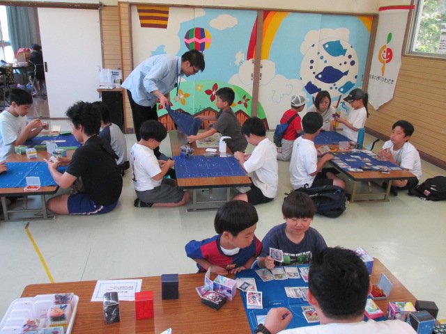 複数のテーブルでポケモンカードをプレイする小中学生や親子で参加しプレイの様子を見守っている写真