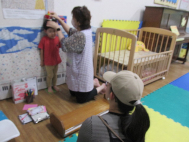 壁に姿勢よく立ち、スタッフに身長を測ってもらっている男の子とそれを見守る保護者の写真
