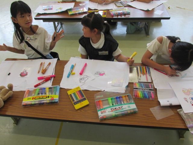 座卓で透明なプラ板にペンや色鉛筆で絵を描く小学生女子3人が並んでいる写真