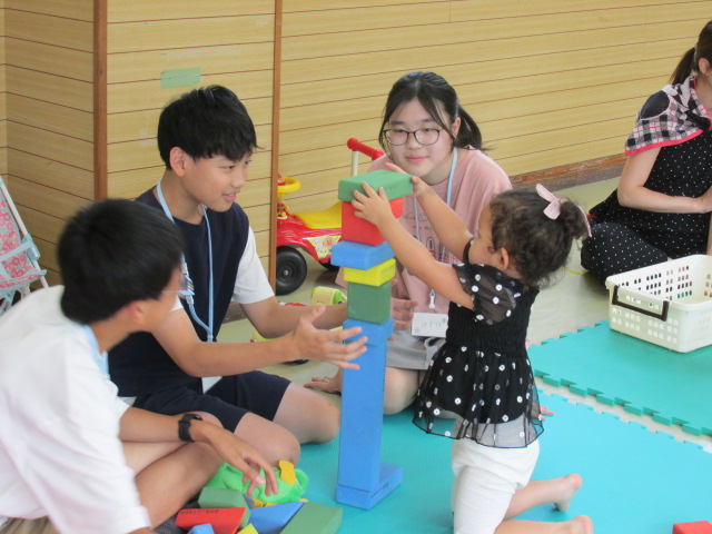 ボランティアに参加した学生3人と幼児が積み木を積み重ねて遊んでいる写真