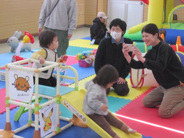 お母さんがすべり台で遊んでいる子どもの写真を撮っている様子