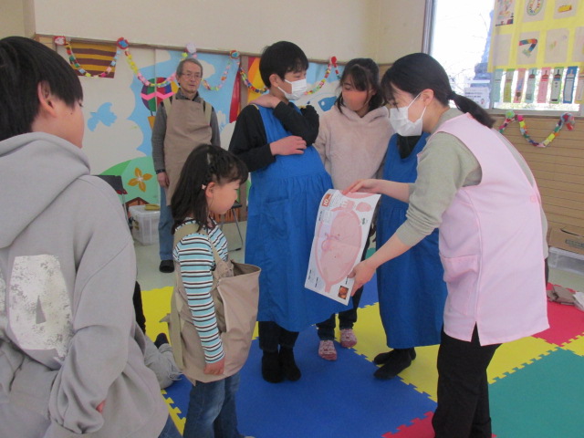 妊婦体験教材を身に着け助産師さんの説明を聞いている子どもたちの写真