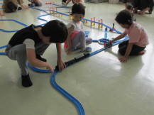 作ったレールの上でプラレールの車両を持ち試走させている2人の子供と間に座りその様子を見る男の子の写真