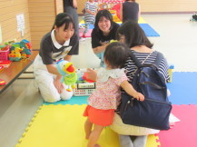 お母さんのリュックサックにしがみついている女の子に中学生の女の子がおもちゃをつかい話しかけている写真