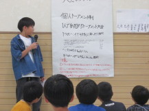 掲示されたルールの紙を見ながら男の子がマイクを持って説明をしている写真