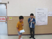 男の子がマイクと資料を持って話をしているそばで1人の男の子が賞状を持って満面の笑顔になっている写真