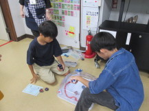 2人の男の子がベイブレードの台の前に対面に座り台の上で回っているお互いのベイブレードを見つめている写真