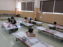 等間隔に並べられた長テーブルの上に新聞紙を敷き複数の子どもたちが白い画用紙に絵を描いている写真